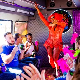 Drag performer in vibrant red costume and wig entertains a cheering, diverse group of passengers on a colorful party bus, guests clapping and holding drinks and pink feather boas.