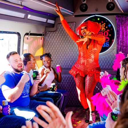 Drag performer in vibrant red costume and wig entertains a cheering, diverse group of passengers on a colorful party bus, guests clapping and holding drinks and pink feather boas.