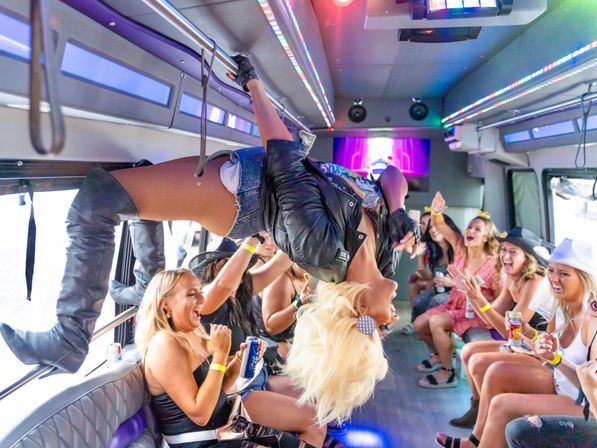 Drag performer hanging upside-down from a handrail entertaining a laughing bachelorette party inside a LED-lit party bus.