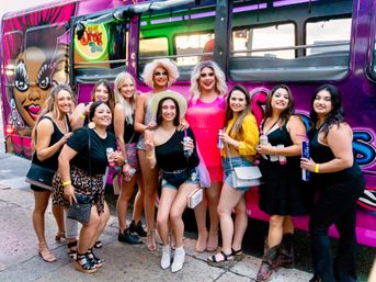 Smiling group of friends with two drag performers posing in colorful outfits in front of a purple party bus mural on a city street, holding drinks for a lively night out.