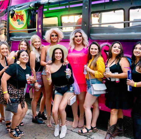 Group of friends posing with two drag performers in bright wigs and fringe dresses outside a pink party bus, holding canned drinks — lively outdoor nightlife scene.