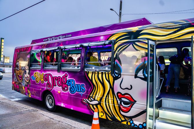Purple pop-art party bus with large drag queen murals, open door revealing passengers and interior lights on an urban street at dusk