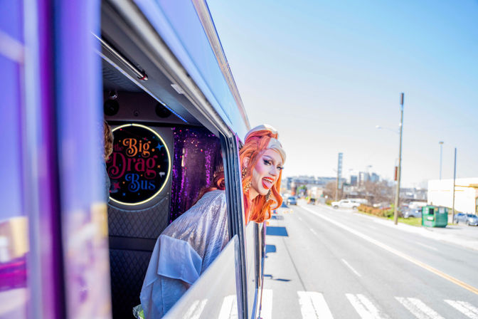 Drag performer with bright orange hair and glittery costume leans out of a purple party bus window, smiling on a sunny urban street.