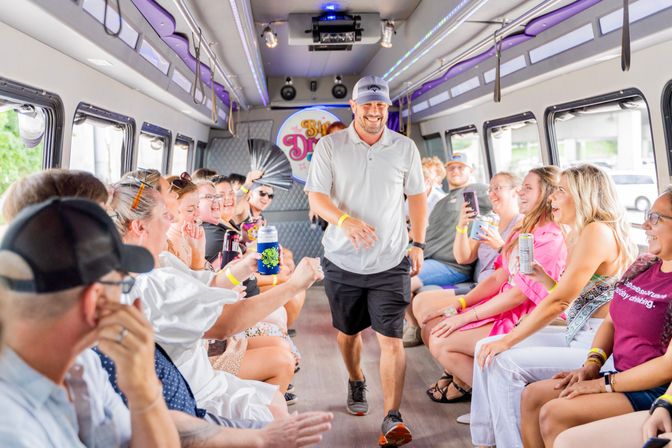 Party bus interior with smiling adults seated along both sides, a man walking down the aisle amid LED lights, drinks and phones during a daytime group celebration.