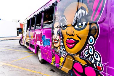 Vibrant purple party bus painted with pop-art drag queen faces, bold makeup and oversized earrings, parked in an urban parking lot
