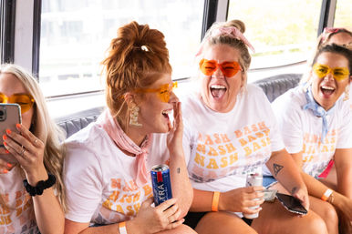 Group of friends in matching 'Bash' t-shirts and colorful sunglasses laughing on a party bus, holding canned drinks, phones, and wearing bandanas.