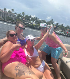 Four friends in colorful swimsuits and heart-shaped sunglasses enjoying drinks on a pontoon boat along a palm-lined waterfront canal under a sunny sky