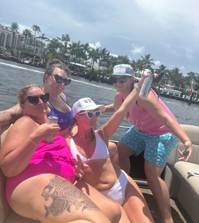 Four friends in colorful swimsuits and heart-shaped sunglasses enjoying drinks on a pontoon boat along a palm-lined waterfront canal under a sunny sky