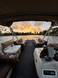 Pontoon boat interior with brown bench seats framing a golden sunset over a palm-lined waterfront and distant city skyline