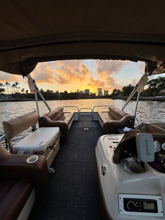 Pontoon boat interior with brown bench seats framing a golden sunset over a palm-lined waterfront and distant city skyline