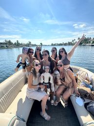 Group of women on a pontoon boat by a palm-lined waterfront canal, enjoying a sunny bachelorette party with inflatable 'groom' dolls and face cutouts.