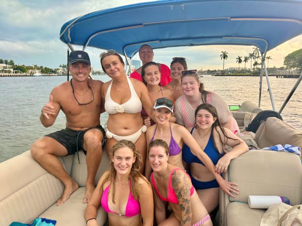 Smiling group of friends in swimwear on a covered pontoon boat cruising a palm-lined tropical waterfront at sunset