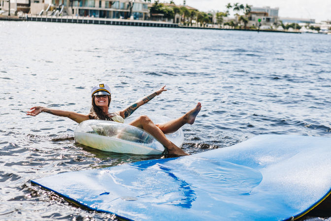 Smiling woman wearing a captain's hat lounging on a clear inflatable tube in calm waterfront waters beside a blue floating mat, coastal homes in the background.