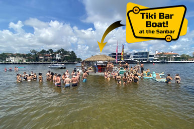 Floating tiki bar boat surrounded by people wading and lounging in shallow water on a sunny day in a residential waterfront inlet with palm trees and luxury homes in the background — lively boating party scene.