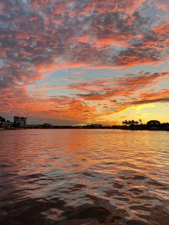Vibrant orange-pink sunset over a tranquil waterfront bay, dramatic cloudscape reflecting on rippling water with silhouetted palm trees and distant shoreline