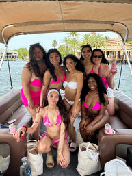 Seven friends in bright pink and white bikinis smiling on a covered pontoon boat, tropical waterfront with palm trees and waterfront homes in the background.