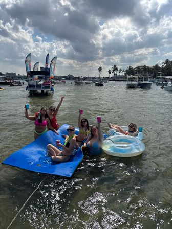Group of friends enjoying a boat party in a sunny coastal bay with palm trees, relaxing on a blue floating mat and clear inner tubes with colorful cups, anchored boats and dramatic clouds in the background.