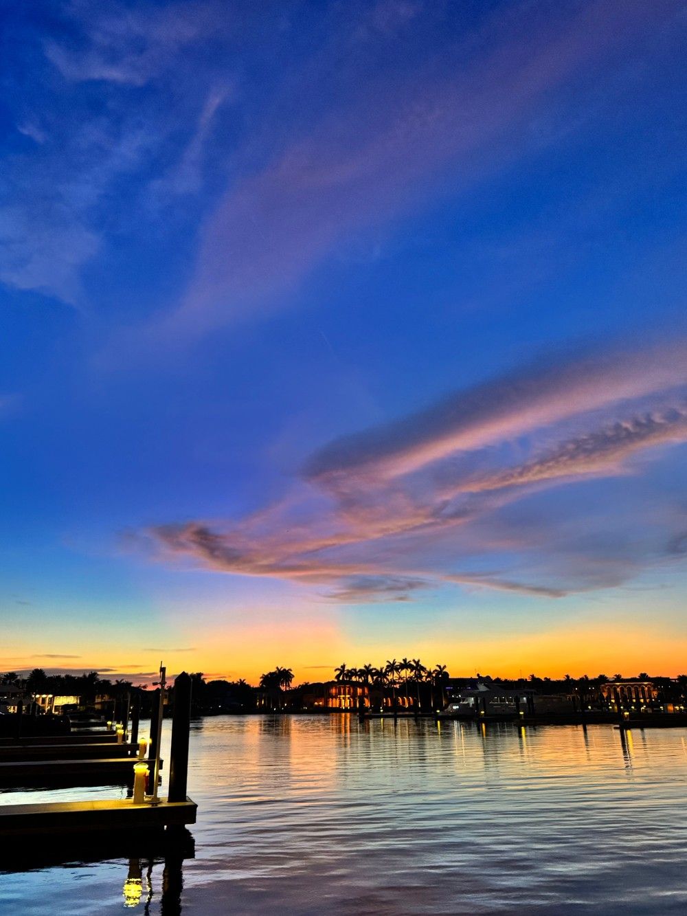 Vibrant sunset over a coastal marina with silhouetted palm trees, glowing dock lights and colorful clouds reflecting on calm water