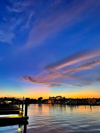 Vibrant sunset over a coastal marina with silhouetted palm trees, glowing dock lights and colorful clouds reflecting on calm water