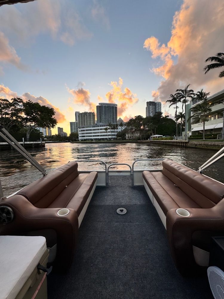 Sunset cruise view from a boat deck with brown bench seats, calm canal water reflecting orange clouds, palm trees and a city skyline of high-rise buildings