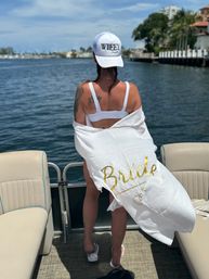 Rear view of a woman in a white bikini and WIFEY cap standing on a pontoon boat, wrapped in a white cover-up with gold "Bride" lettering, overlooking blue waterfront and marina homes.