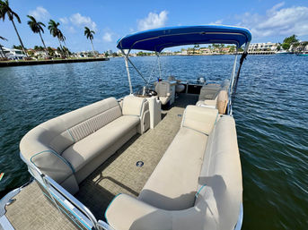 Pontoon boat with tan cushioned lounges and blue bimini cruising a palm-lined coastal waterway past waterfront homes under a sunny blue sky