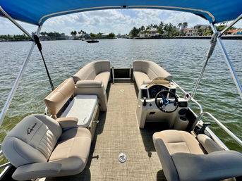 View from a pontoon boat with tan cushioned seating and captain's console under a blue bimini, cruising on calm water past palm-lined waterfront homes on a sunny day.
