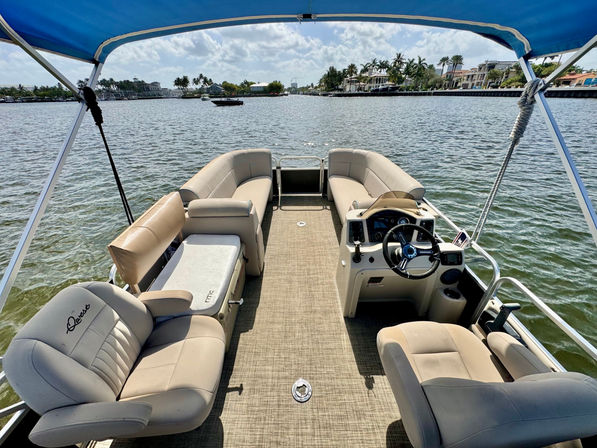View from a pontoon boat with tan cushioned seating and captain's console under a blue bimini, cruising on calm water past palm-lined waterfront homes on a sunny day.