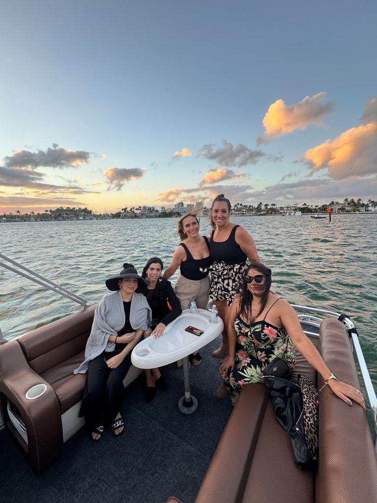 Five women smiling on a pontoon boat during a sunset cruise, palm-lined waterfront skyline and colorful clouds over calm water.