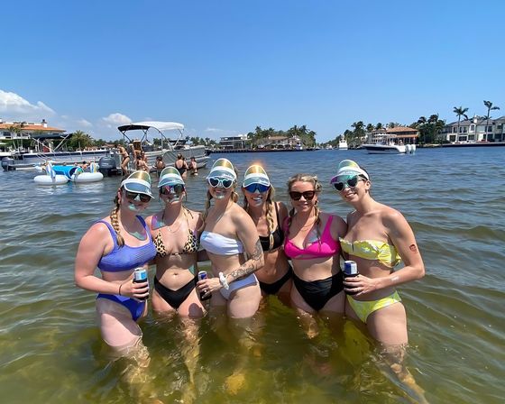 Six friends in colorful bikinis standing waist-deep in a sunny coastal waterway holding drinks, surrounded by boats, inflatable floats, palm trees and waterfront homes