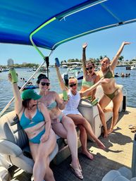 Five friends in swimsuits cheering with canned drinks on a pontoon boat under a blue canopy at a sunny marina with palm trees and other boats nearby.