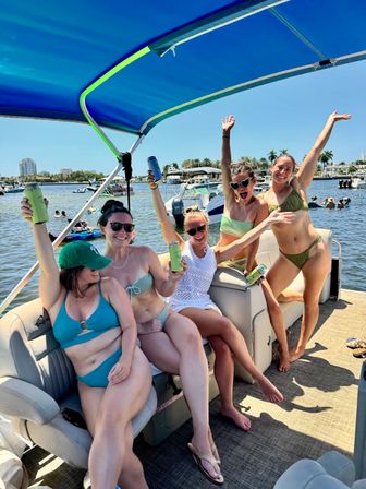 Five friends in swimsuits cheering with canned drinks on a pontoon boat under a blue canopy at a sunny marina with palm trees and other boats nearby.