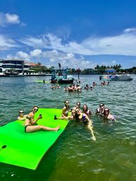 Group of friends enjoying a sunny boat party in a calm coastal inlet, lounging on a bright green floating mat and wading near anchored boats and waterfront homes under a blue sky.