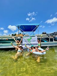 Five friends in swimsuits cheering in shallow green water beside a blue-canopied pontoon boat, holding drinks and a 'MARGS & MATRIMONY EST. 2025' banner under a bright sunny sky.