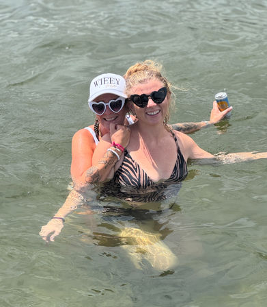 Two smiling women waist-deep in shallow beach water on a sunny summer day; one wears a white cap with 'WIFEY' text and white heart-shaped sunglasses, the other wears a zebra-print swimsuit and black heart sunglasses while holding a canned drink.