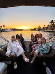 Group of women enjoying a sunset pontoon boat cruise on a palm-lined waterfront canal with yachts, luxury homes, and drinks
