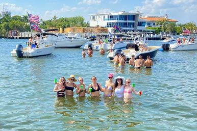 Sunny boat-party scene with a group of friends in swimsuits wading waist-deep in a shallow bay holding colorful cups, surrounded by anchored recreational boats, palm-lined waterfront homes, and a bright blue sky.