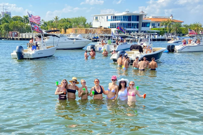 Sunny boat-party scene with a group of friends in swimsuits wading waist-deep in a shallow bay holding colorful cups, surrounded by anchored recreational boats, palm-lined waterfront homes, and a bright blue sky.