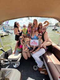 Group of friends in swimsuits posing on a sunny pontoon boat at a marina with palm trees and boats in the background, one holding a playful sign reading “NO WEENIES ALLOWED”.