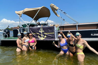 Six women in colorful bikinis and mirrored visors posing waist-deep in shallow green water next to a tan-canopied pontoon boat, smiling and holding drinks on a sunny waterfront day.