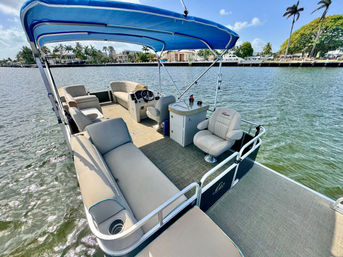Pontoon boat interior with beige cushioned lounges, swivel captain’s chair and helm under a bright blue bimini top, cruising a sunny waterfront canal lined with palm trees and waterfront homes.