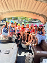 Eight friends in patriotic USA shirts and neon caps smiling aboard a covered pontoon boat on a sunny waterfront canal.