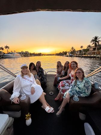 Friends enjoying a sunset boat cruise on a palm‑lined waterfront canal with yachts and a golden sky