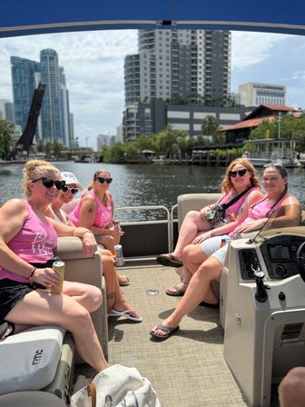 Five women in matching pink tank tops relaxing on a pontoon boat during a sunny river cruise past a downtown waterfront skyline