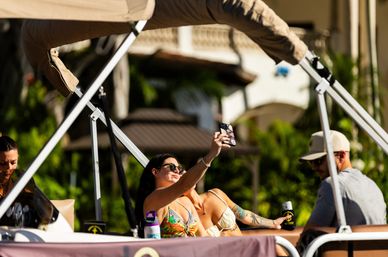 Friends lounging on a pontoon boat in bright sunlight — a woman in a colorful bikini taking a selfie while companions relax with drinks against a tropical resort backdrop.