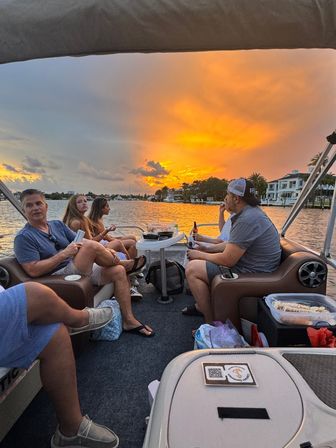 Group of people lounging on a pontoon boat during a vibrant orange sunset, calm water reflecting the sky and waterfront homes along the shoreline — relaxed sunset boat ride.