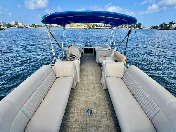 Spacious pontoon boat deck with beige cushioned benches and blue bimini top, floating on calm blue water with waterfront homes and a sunny sky