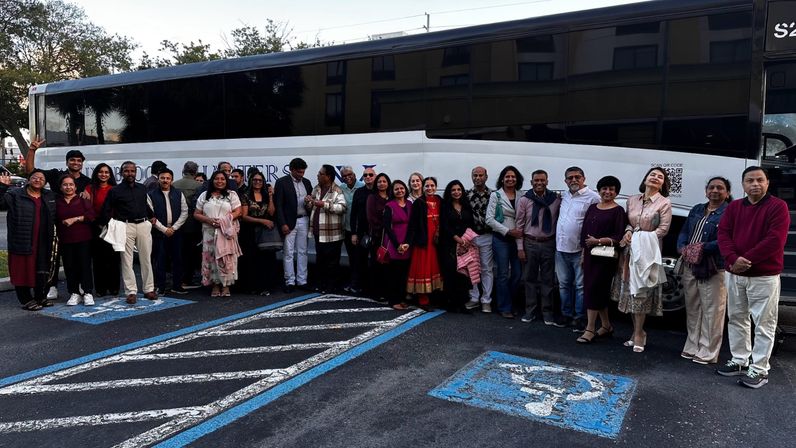 Large group of adults smiling and posing in front of a white charter coach bus in a hotel parking lot, standing across accessible parking spaces and striped pavement.