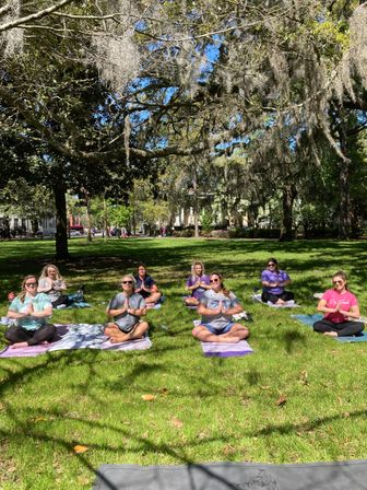 Group doing outdoor yoga on mats in a sunny park, meditating beneath Spanish-moss‑draped oak trees