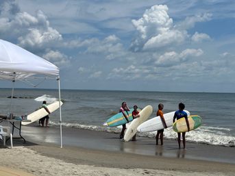 Group of surfers with colorful surfboards standing at a sandy beach shoreline next to a white canopy tent, facing gentle ocean waves under a partly cloudy sky